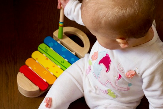 A baby is sitting on a wooden floor, wearing a white outfit with colorful patterns. In front of the baby is a rainbow-colored toy xylophone. The baby holds a mallet and appears to be playing with the xylophone.