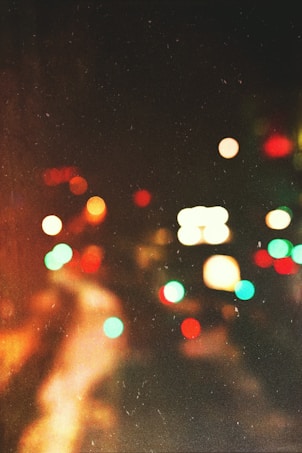A soft-focus image with blurred colorful lights against a dark background, evoking a sense of a city street at night. The lights are in hues of red, green, and orange, resembling distant car lights or streetlights.