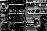 A collection of vintage cameras and photography equipment is neatly arranged on shelves. Various models of cameras, lenses, and accessories are displayed, alongside a selection of watches in a display case. The setting has a retro and nostalgic feel, likely in a shop or exhibition.