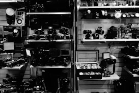 A collection of vintage cameras and photography equipment is neatly arranged on shelves. Various models of cameras, lenses, and accessories are displayed, alongside a selection of watches in a display case. The setting has a retro and nostalgic feel, likely in a shop or exhibition.