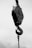A black and white image of a heavy-duty metal crane hook suspended from a cable, showing wear and exposed to the elements. The intricate mechanism and rivets are prominently featured, with a cloudy sky backdrop.