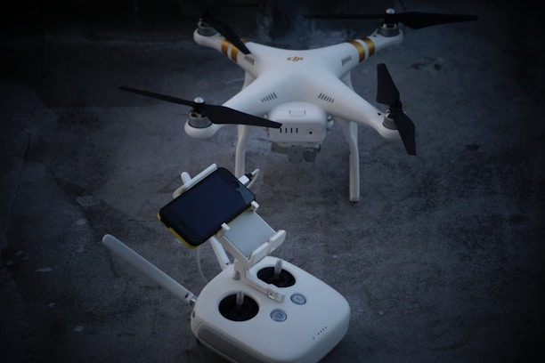 A white quadcopter drone with four rotors is resting on a concrete surface. Accompanying the drone is a white remote controller with dual joysticks and a smartphone mounted on the top.