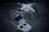 A white quadcopter drone with four rotors is resting on a concrete surface. Accompanying the drone is a white remote controller with dual joysticks and a smartphone mounted on the top.