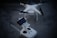 A white quadcopter drone with four rotors is resting on a concrete surface. Accompanying the drone is a white remote controller with dual joysticks and a smartphone mounted on the top.