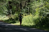 A person is running along a shaded path surrounded by dense, lush green foliage and trees. The sunlight filters through the canopy, casting dappled shadows on the ground. The runner is wearing athletic gear, including a hat and brightly colored socks.