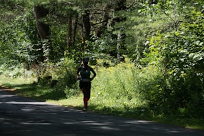 A person is running along a shaded path surrounded by dense, lush green foliage and trees. The sunlight filters through the canopy, casting dappled shadows on the ground. The runner is wearing athletic gear, including a hat and brightly colored socks.