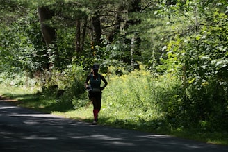 A person is running along a shaded path surrounded by dense, lush green foliage and trees. The sunlight filters through the canopy, casting dappled shadows on the ground. The runner is wearing athletic gear, including a hat and brightly colored socks.