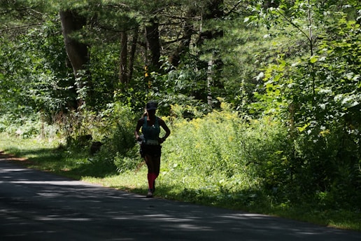 A person is running along a shaded path surrounded by dense, lush green foliage and trees. The sunlight filters through the canopy, casting dappled shadows on the ground. The runner is wearing athletic gear, including a hat and brightly colored socks.