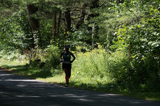 A person is running along a shaded path surrounded by dense, lush green foliage and trees. The sunlight filters through the canopy, casting dappled shadows on the ground. The runner is wearing athletic gear, including a hat and brightly colored socks.