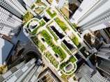 A top-down view of a modern urban landscape featuring tall, symmetrical buildings surrounding a central area filled with greenery. The scene includes rooftops with gardens, structured pathways, and recreational spaces, highlighting the integration of nature within the architectural framework. The geometric design and layout of the structures and greenery create a visually appealing, organized aesthetic.