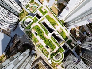 A top-down view of a modern urban landscape featuring tall, symmetrical buildings surrounding a central area filled with greenery. The scene includes rooftops with gardens, structured pathways, and recreational spaces, highlighting the integration of nature within the architectural framework. The geometric design and layout of the structures and greenery create a visually appealing, organized aesthetic.