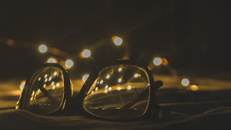 A pair of eyeglasses is placed on a surface with a background of blurred, warmly lit fairy lights. The glow from the lights reflects off the lenses, creating a cozy and inviting ambiance.