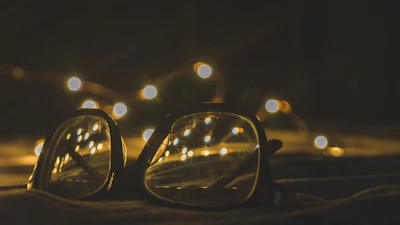 A pair of eyeglasses is placed on a surface with a background of blurred, warmly lit fairy lights. The glow from the lights reflects off the lenses, creating a cozy and inviting ambiance.