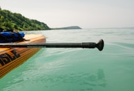 A paddle board with wooden stripes is floating on clear turquoise water, close to the shore. A paddle is resting on the board, and a lush green hillside can be seen in the background under a soft, pale sky.