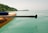 A paddle board with wooden stripes is floating on clear turquoise water, close to the shore. A paddle is resting on the board, and a lush green hillside can be seen in the background under a soft, pale sky.