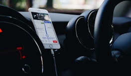 A smartphone is mounted on a car dashboard displaying a navigation app, showing directions and a map. The interior of the car is visible, including the steering wheel and air vents.