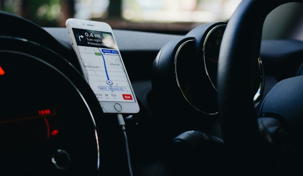 A smartphone is mounted on a car dashboard displaying a navigation app, showing directions and a map. The interior of the car is visible, including the steering wheel and air vents.