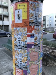 A cylindrical poster column covered with various colorful posters. The posters advertise events, art shows, and markets, with prominent use of text and diverse graphic designs. The column is situated on a street with nearby buildings and a parked car visible in the background.