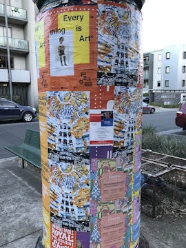 A cylindrical poster column covered with various colorful posters. The posters advertise events, art shows, and markets, with prominent use of text and diverse graphic designs. The column is situated on a street with nearby buildings and a parked car visible in the background.