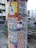 A cylindrical poster column covered with various colorful posters. The posters advertise events, art shows, and markets, with prominent use of text and diverse graphic designs. The column is situated on a street with nearby buildings and a parked car visible in the background.
