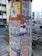 A cylindrical poster column covered with various colorful posters. The posters advertise events, art shows, and markets, with prominent use of text and diverse graphic designs. The column is situated on a street with nearby buildings and a parked car visible in the background.