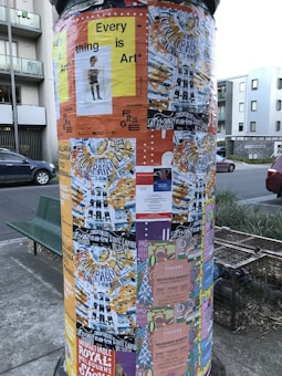 A cylindrical poster column covered with various colorful posters. The posters advertise events, art shows, and markets, with prominent use of text and diverse graphic designs. The column is situated on a street with nearby buildings and a parked car visible in the background.