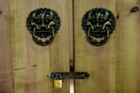 Wooden doors feature ornate metal lion head knockers with rings and a small brass padlock securing a horizontal latch. The wood has a natural, rustic appearance with visible grain.