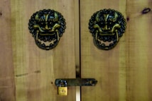 Wooden doors feature ornate metal lion head knockers with rings and a small brass padlock securing a horizontal latch. The wood has a natural, rustic appearance with visible grain.