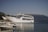 A large white ferry named 'WESTFERRY' with 'CAPTAIN ARISTIDIS' written on its side is docked next to a pier. Smaller boats are moored in the foreground, and a mountainous landscape with sparse vegetation is visible in the background under a clear blue sky.
