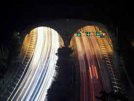 A night scene featuring a multi-lane highway tunnel with bright light streaks indicating moving vehicles. The left lane has white and yellow light trails while the right lane has mainly red light trails, suggesting incoming and outgoing traffic. Overhead traffic signals with green arrows direct the flow. Trees and side barriers are vaguely visible along the road.