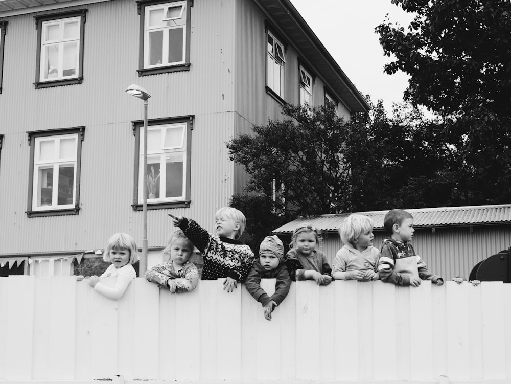 grayscale photo of seven children standing in wooden fence near house