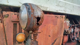 A close-up view of a rusty, damaged headlight on an old vehicle. The metal casing is corroded with holes, exposing deterioration over time. The headlight is attached to a rusty, orange panel with visible bolts and wires. The background includes intricate metal components and faded white paint.