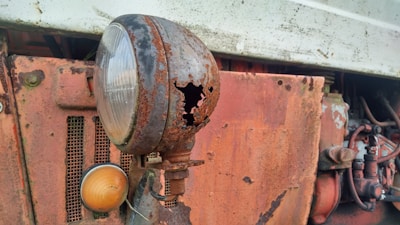 A close-up view of a rusty, damaged headlight on an old vehicle. The metal casing is corroded with holes, exposing deterioration over time. The headlight is attached to a rusty, orange panel with visible bolts and wires. The background includes intricate metal components and faded white paint.