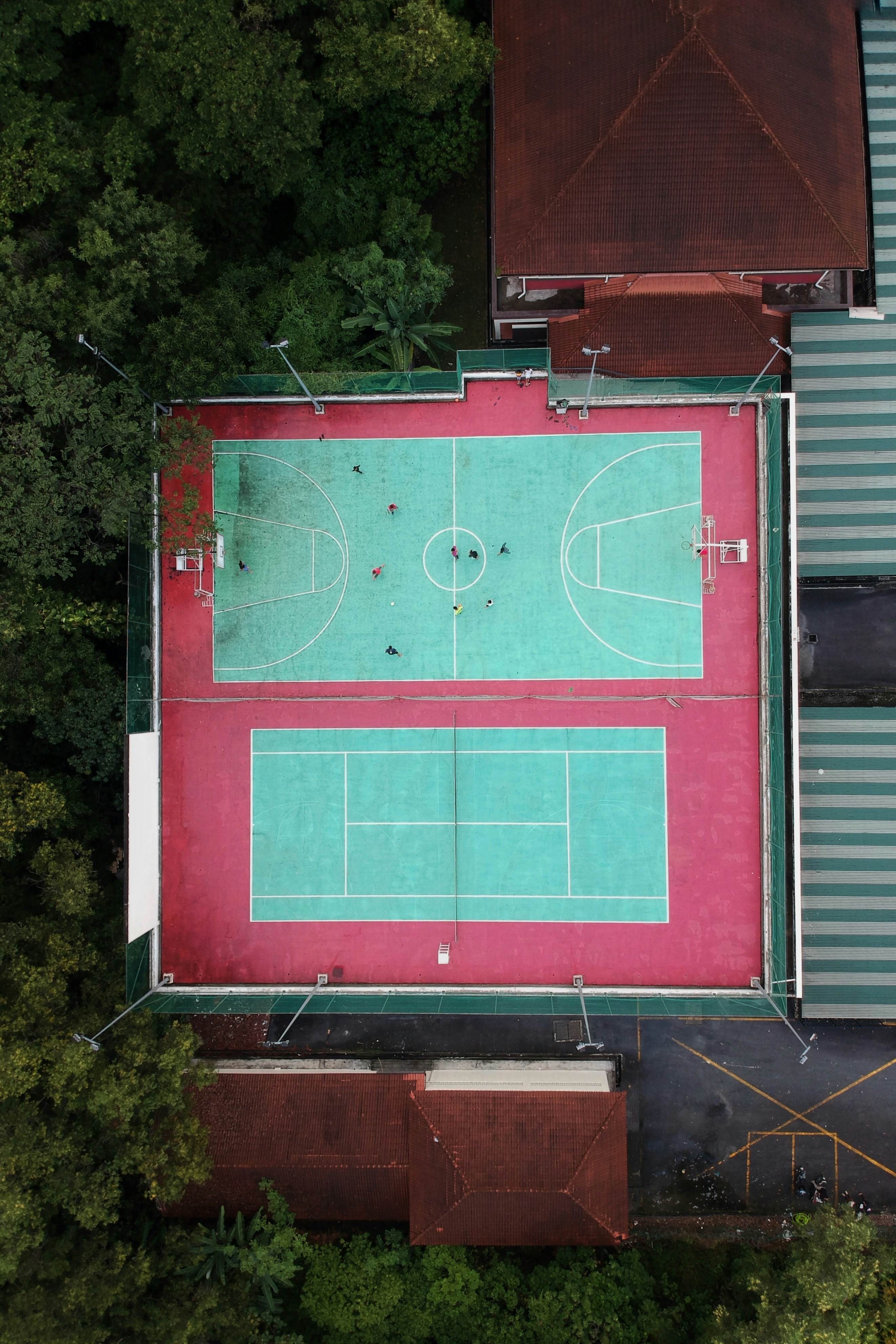 The aerial view depicts a multi-sport court featuring both a basketball court and a tennis court, surrounded by lush green trees and adjacent buildings. Players are visible on the basketball court, engaging in a game. The court's surface is primarily teal with red borders, and there are overhead light fixtures around the perimeter.