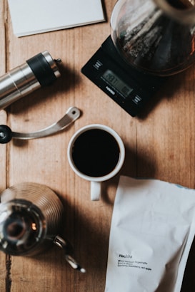 A coffee brewing setup is arranged on a wooden surface, including a white cup of black coffee, a silver manual coffee grinder, a stainless steel kettle, a digital scale displaying 437.7 grams, a coffee dripper with a brownish liquid inside, and a white bag of coffee labeled 'Hachira'.