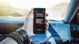 A hand holding a smartphone displaying a motivational message on the screen while inside a car. The phone screen reads 'How bad do you want it?' The hand is wearing several bracelets and a watch. In the background, a car interior and a laptop with a mountain wallpaper are visible.