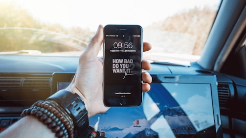A hand holding a smartphone displaying a motivational message on the screen while inside a car. The phone screen reads 'How bad do you want it?' The hand is wearing several bracelets and a watch. In the background, a car interior and a laptop with a mountain wallpaper are visible.
