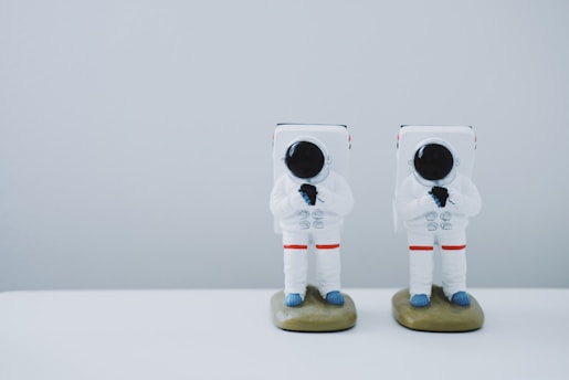 Two toy astronauts with reflective black visors and white suits stand side by side on a light surface. Each astronaut features blue gloves and footwear, with red accents on their suit. They are positioned against a plain light gray background.