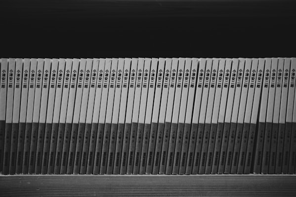 A row of identically bound books with Japanese text on the spines, arranged neatly side by side on a wooden shelf. The image is in black and white, emphasizing the uniformity and orderly presentation of the books.