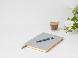 A minimalist setting featuring an elegant notebook with a pen resting on it. The notebook has a gray cover with gold edges and a brown ribbon bookmark. A small glass of coffee sits nearby alongside blooming white flowers and green leaves, all set against a clean white background.
