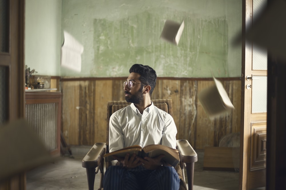 homme assis sur un fauteuil en bois brun tout en portant un livre brun