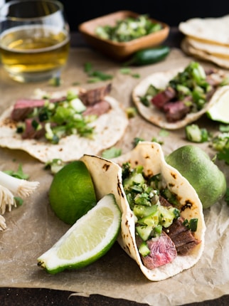 Soft tacos filled with grilled steak and topped with diced green vegetables and herbs rest on parchment paper. A slice of lime is placed beside the tacos, along with whole limes and a glass of light-colored beverage in the background. A wooden bowl holding more green vegetables and a stack of tortillas are visible.