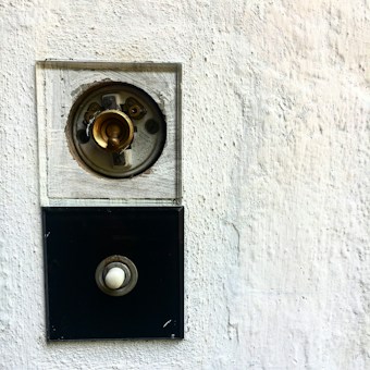 A close-up of a vintage light switch installed on a textured white wall. The switch has a round brass component at the top with visible screws and wiring. Below, there is a black square plate with a white circular button.