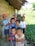 A group of young children are gathered outside a rustic wooden structure with a makeshift thatched roof. They are standing on a narrow path beside a lush green field. Some children are dressed in colorful shirts, and one boy is wearing a bright orange cap. The mood appears lighthearted and playful, with some children smiling and others looking curiously at the camera.
