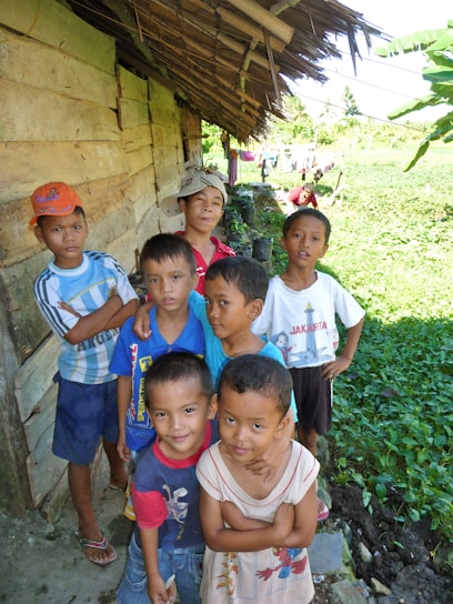 A group of young children are gathered outside a rustic wooden structure with a makeshift thatched roof. They are standing on a narrow path beside a lush green field. Some children are dressed in colorful shirts, and one boy is wearing a bright orange cap. The mood appears lighthearted and playful, with some children smiling and others looking curiously at the camera.