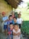 A group of young children are gathered outside a rustic wooden structure with a makeshift thatched roof. They are standing on a narrow path beside a lush green field. Some children are dressed in colorful shirts, and one boy is wearing a bright orange cap. The mood appears lighthearted and playful, with some children smiling and others looking curiously at the camera.