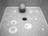 A human skull placed in a tiled shower corner with circular patterns on the floor and a central drain. The tiles are arranged in a geometric pattern.