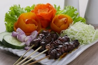 A decorative plate featuring grilled meat skewers accompanied by fresh vegetables. The skewers have a glossy glaze, while the plate is garnished with intricately carved orange vegetable flowers, finely shredded cabbage, thinly sliced red onions, and cucumber slices, all presented on a bed of leafy greens.