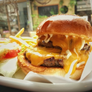 A juicy cheeseburger with melted cheese and layers of beef patty sits on a plate. Next to it is a portion of golden French fries and a salad with lettuce and tomato. The background is slightly blurred, suggesting an indoor setting with natural light.