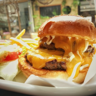 A juicy cheeseburger with melted cheese and layers of beef patty sits on a plate. Next to it is a portion of golden French fries and a salad with lettuce and tomato. The background is slightly blurred, suggesting an indoor setting with natural light.